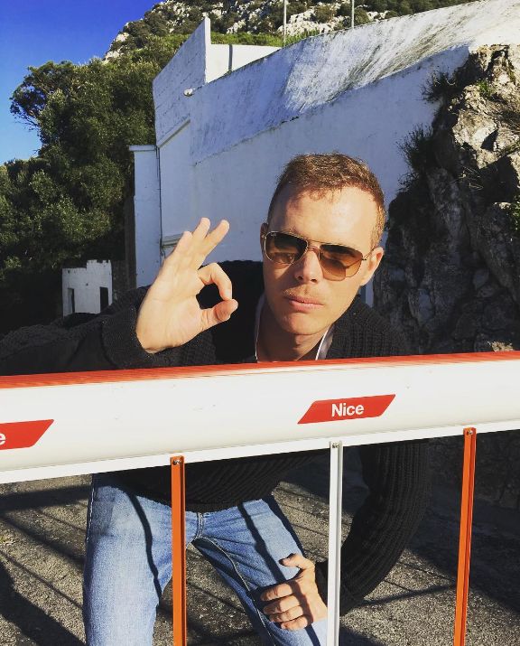 Martyn in aviator sunglasses making an OK sign, leaning over a red and white car park barrier that says Nice.